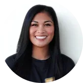 A woman with long black hair and a black shirt stands smiling in front of a plain, light-colored background.