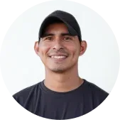 Man wearing a black baseball cap and dark t-shirt, smiling, standing against a plain white background.