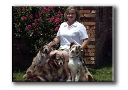A woman sitting outdoors with three dogs in front of a brick wall and flowering bush.