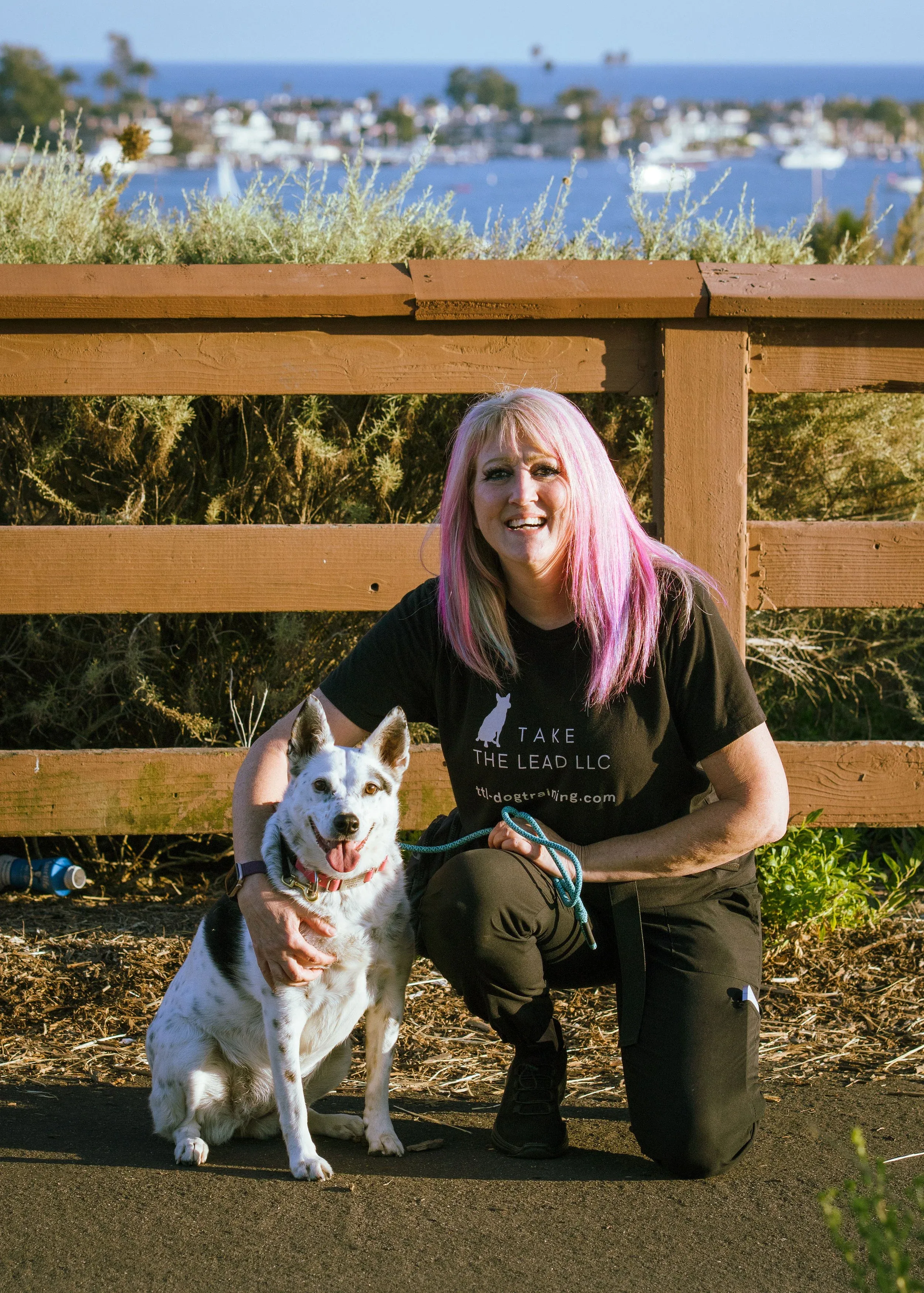 A woman with pink hair smiling and kneeling next to a happy white and black dog outdoors, with a wooden fence, greenery, and a body of water with boats in the background.